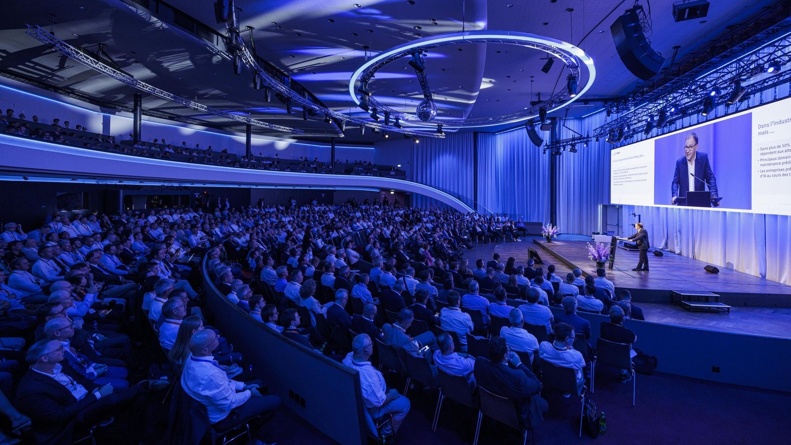 Conference hall with a speaker on stage, a presentation screen and a large seated audience